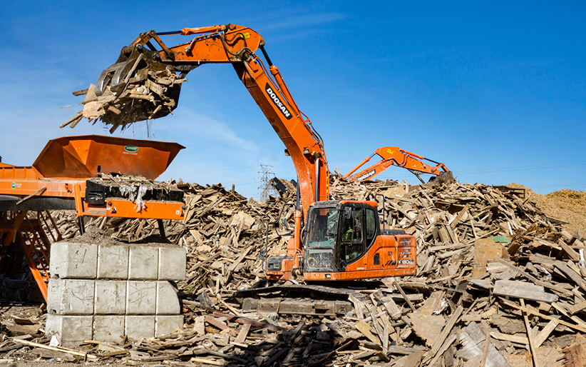 A Doosan DX180LC-3 crawler excavator uses a grapple attachment to place construction debris on a conveyor belt for sorting purposes.