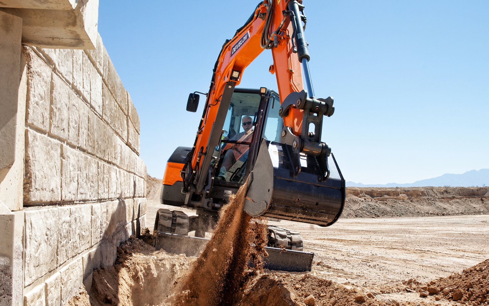A DEVELON compact excavator digs next to a wall.